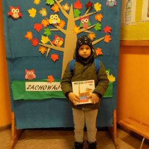 Korytarz w budynku szkoły w Zawadowie. Chłopiec stoi, trzyma książkę i kartę.W tle dekoracja -kolorowe drzewo.
