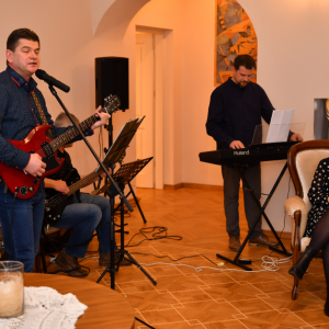 CPT w Ciechankach. Na zdjęciu z lewej strony stoi mężczyzna, grająć na gitarze śpiewa do mikrofonu, z prawej strony w fotelu siedzi kobieta w tle dwaj meżczyźni grający na instrumentach.