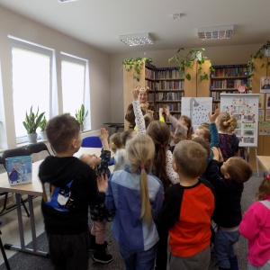 Wnętrze biblioteki. Dzieci stoją w grupie i trzymaja w górze ręce. 